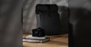 A nikon camera rests on two books beside a closed black backpack against a textured gray background. the setting is simple and stylish, emphasizing the camera and bag.