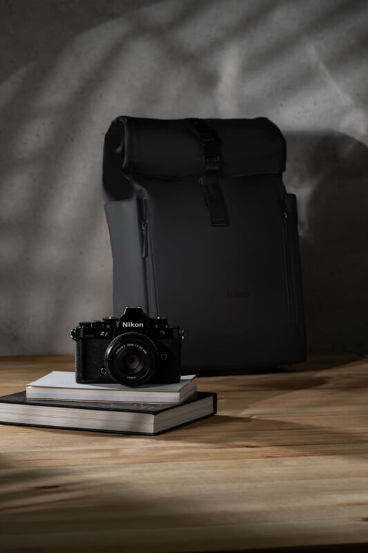 A nikon camera sits on top of a closed book, which rests on a wooden table. next to the camera is a black, upright modern backpack, all under soft, dappled lighting.