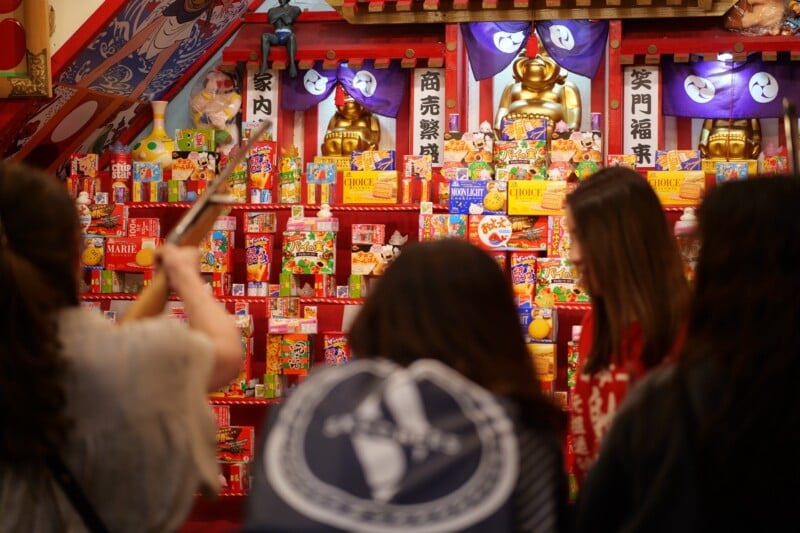 A group of people are participating in a festival game booth decorated with colorful packages and toys. One person aims an air rifle at a prize, attempting to win. The booth features Japanese signs and a golden statue in the background.