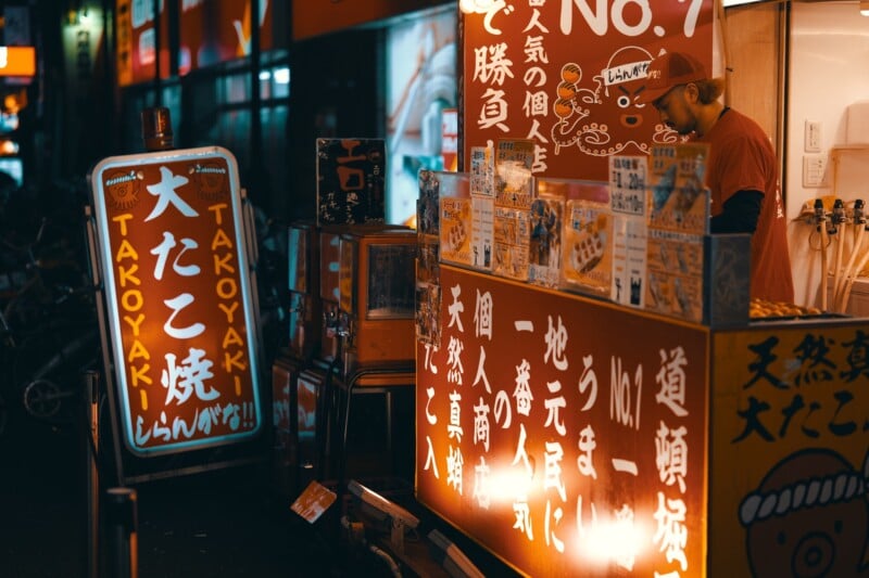A busy street food stall at night in Japan, featuring bright orange signage with Japanese text and a takoyaki sign. A person wearing a hat stands behind the counter preparing food. The warm glow of the lights illuminates the scene.