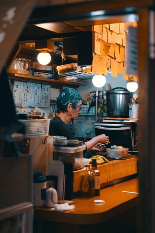 A man wearing a bandana and a black shirt is busy preparing food behind a wooden counter in a cozy, dimly lit restaurant or food stall. Shelves filled with cups, jars, and other kitchen items are seen in the background. Warm lighting enhances the intimate setting.