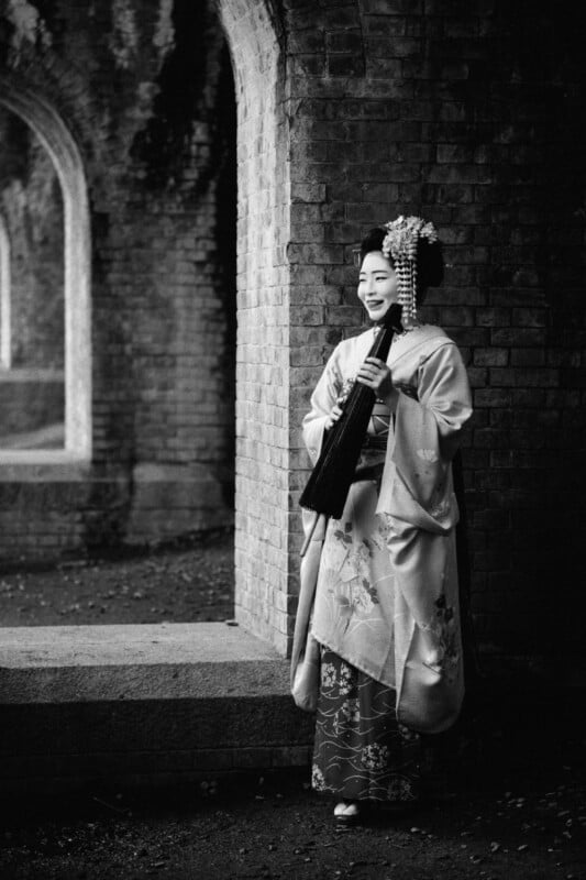 Black and white photo of a person in traditional Japanese attire standing by a brick archway. They are wearing a kimono with intricate designs and holding an umbrella, smiling and gazing to the side. The background includes multiple arches with shadows and texture.