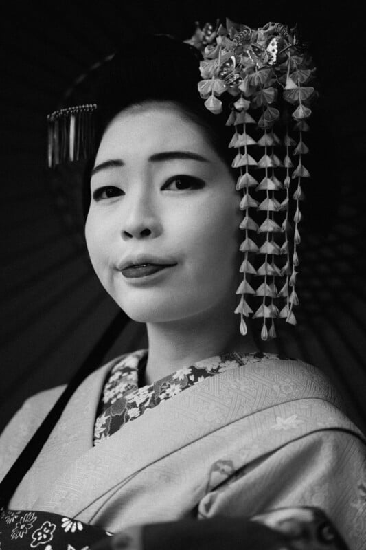 A black and white portrait of a woman dressed in traditional Japanese attire. She is wearing a decorated kimono and an elaborate hairpiece with hanging ornaments. Her expression is calm and poised.