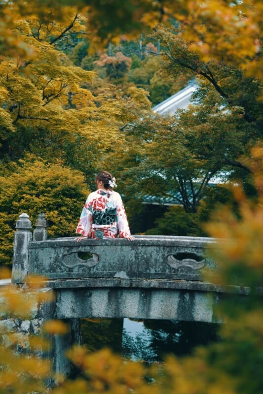 A person wearing a traditional Japanese kimono stands on an old stone bridge, surrounded by lush autumn foliage. The scene is peaceful, with rich green and golden leaves framing the bridge and a glimpse of a building roof in the background.