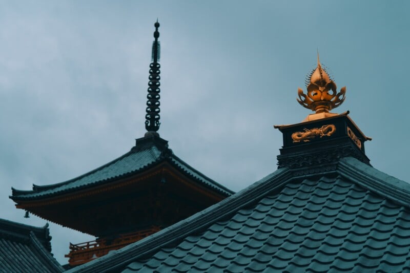 A traditional Japanese temple with intricate tiled rooftops set against a cloudy sky. The structure features ornate details, including a spire and a golden ornamental piece, highlighting the architectural beauty and cultural significance of the temple.