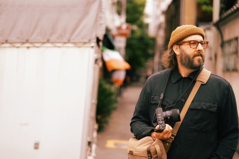 A bearded person wearing a brown beanie, eyeglasses, and a dark jacket stands in an outdoor urban setting holding a camera. They have a tan shoulder bag strapped across their body and appear to be looking thoughtfully to the side with buildings and greenery in the background.