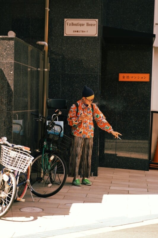 A person wearing a colorful shirt and pants stands on the sidewalk pointing to the right while holding a smartphone. A bicycle is parked nearby against the black building wall reflecting sunlight. There are Japanese signs on the wall and an orange sign in the background.