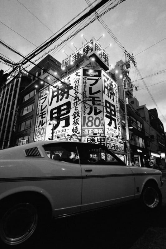 A black and white photo of a bustling city street in Japan during twilight. A vintage car passes by a brightly lit building adorned with large, bold Japanese signs. Overhead wires crisscross above, and several other buildings are visible in the background.