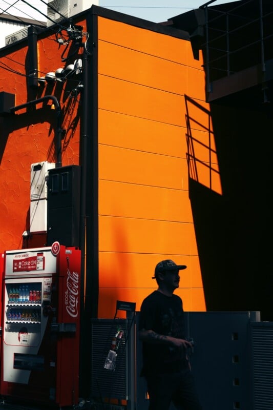A bright orange wall in sunlight, casting sharp shadows including a staircase silhouette. A red vending machine dispensing beverages is positioned near the wall. An individual in a camo hat and dark clothes walks past, carrying a transparent plastic bottle.