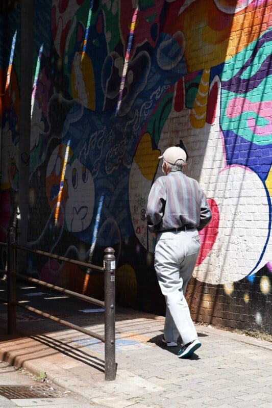 An elderly man wearing a striped shirt and light-colored pants walks by a colorful mural painted on a brick wall. The mural features various abstract shapes and patterns with vibrant colors. The scene is lit by sunlight, casting shadows on the ground.