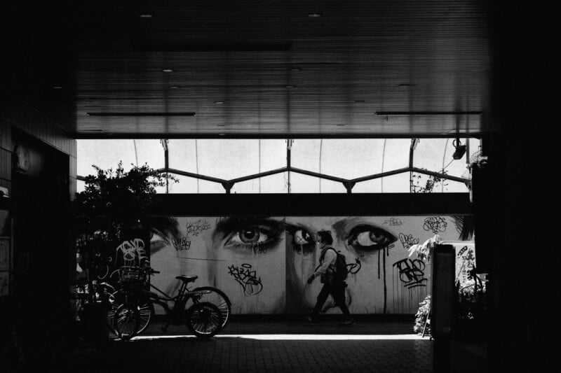 A person walks through a dimly lit tunnel adorned with graffiti, featuring large, intense eyes on the wall. A bicycle is parked on the left side, and sunlight filters through the ceiling onto the pavement below.