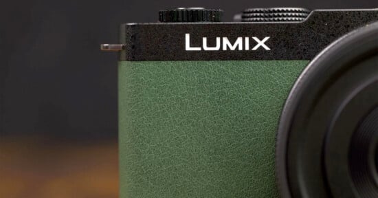 Close-up of a Lumix camera, showing the upper left portion. The camera features a black textured top with a black dial and a green textured grip. Part of the lens is visible on the right side of the image. The brand name "Lumix" is prominently displayed.