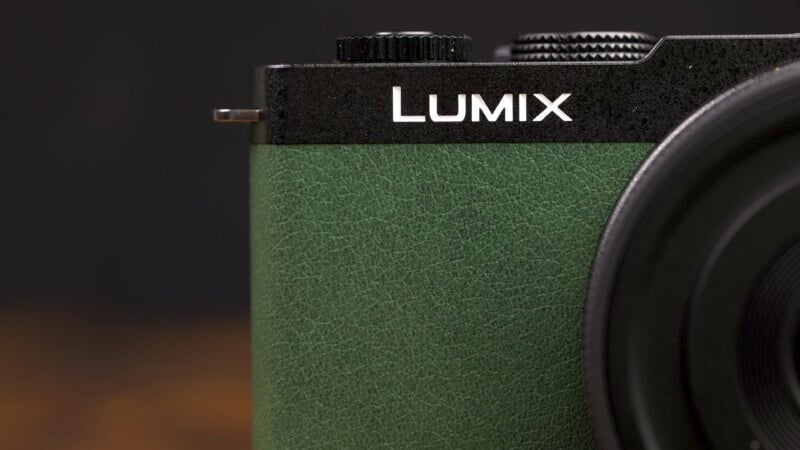 Close-up image of a green and black Lumix camera with textured surface and a silver button on the top. The photo captures part of the lens and the Lumix brand name in clear white letters on the top front edge.