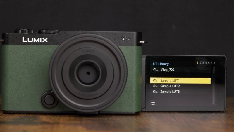 A Lumix camera with a green body is displayed on a wooden surface. To the right of the camera is an extended screen showing a LUT (Lookup Table) library menu with options including "Vlog_709" and "Sample LUT1, LUT2, LUT3.