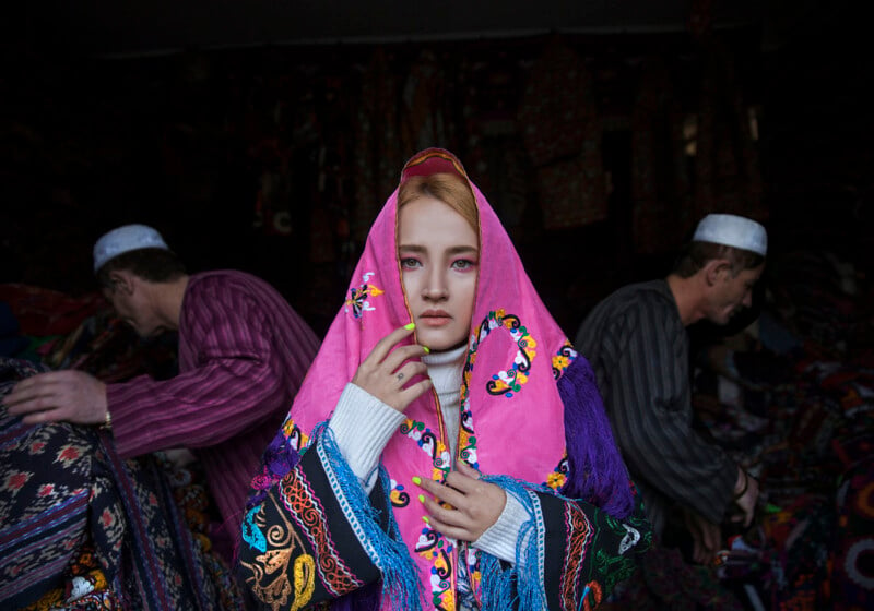 A young woman wearing a vibrant, patterned pink headscarf stands in the foreground with a thoughtful expression. Two men in traditional attire are seen in the background, busy with textiles in a dimly lit room filled with colorful fabric.