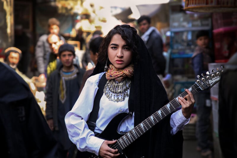 A woman dressed in a white blouse, colorful scarf, and traditional jewelry holds an electric guitar. She stands in a crowded marketplace, surrounded by onlookers. The setting appears vibrant and busy, with various stalls and people in the background.