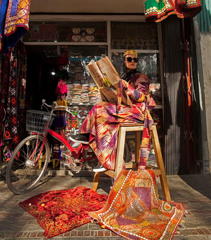 A person dressed in colorful, intricate clothing and wearing sunglasses and a decorative headpiece sits on a stool outside a vibrant textiles shop while holding an open book. A bicycle rests nearby, and elaborate fabrics are displayed on the ground and hanging around.