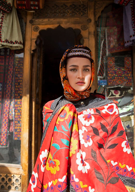 A woman stands in front of a shop entrance, adorned in a colorful headscarf and shawl featuring intricate floral patterns. The background showcases traditional textile artworks and wooden door frames, highlighting cultural craftsmanship.