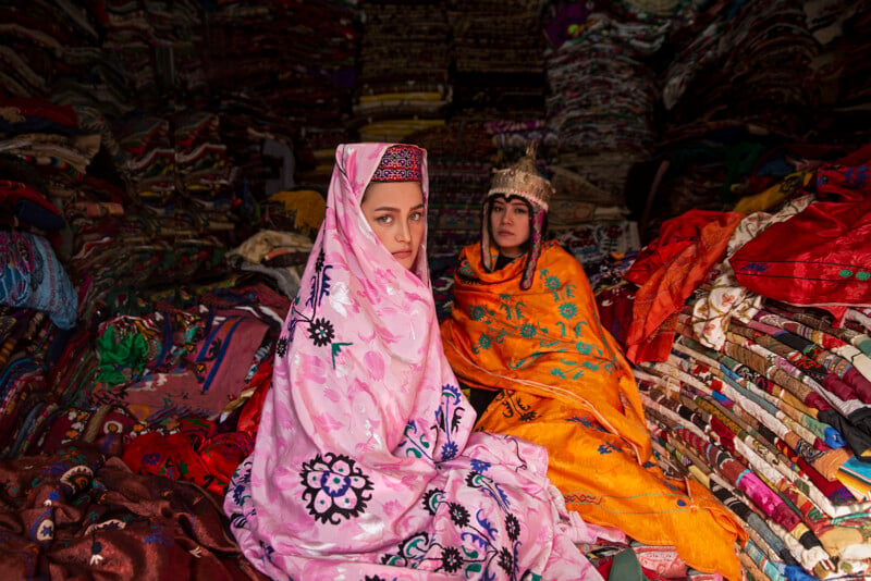 Two women are sitting amidst a vibrant array of textiles in a market. One woman wears a pink shawl with black and purple floral patterns, while the other wears an orange shawl with blue floral designs and a decorative headdress. Shelves filled with folded fabrics surround them.