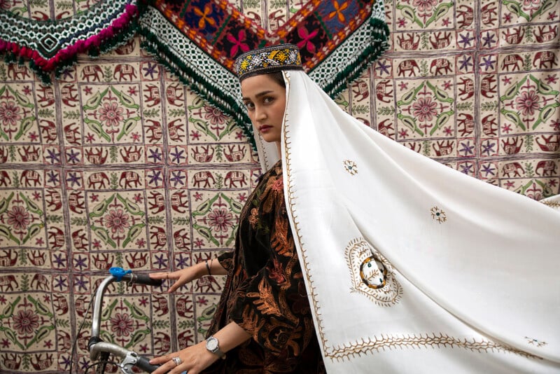 A woman in traditional attire with a white, embroidered headscarf stands next to a bicycle. She wears an ornate cap and a patterned robe. Behind her, there are intricately designed textiles with elephants and floral motifs hanging as a backdrop.