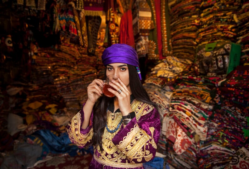 A person wearing a vibrant traditional outfit and a purple headscarf is drinking tea in a richly decorated room filled with colorful textiles and fabrics stacked on shelves. The scene is lively with a variety of intricate patterns and textures.