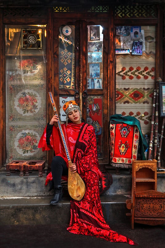 A person dressed in vibrant traditional attire, adorned with a striking red and black patterned garment and headpiece, sits in front of a shop adorned with colorful textiles and decorations. They hold a long stringed musical instrument, adding to the cultural ambiance.