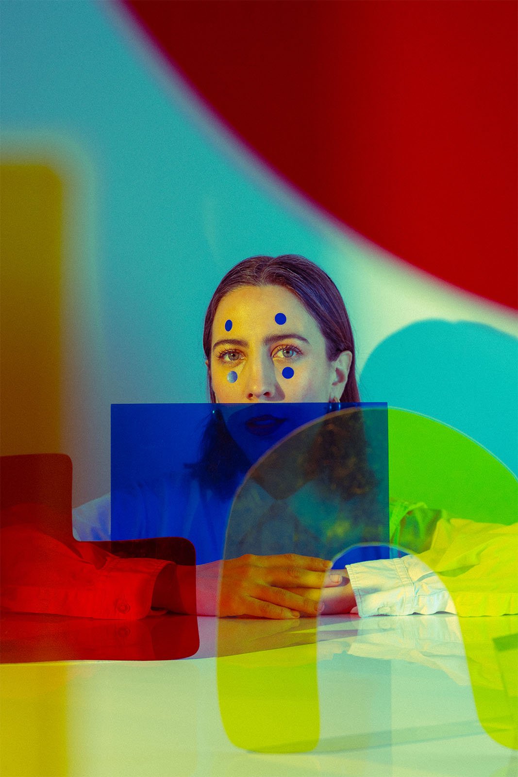 A person with blue dots on their face gazes directly at the camera. They are seated behind a colorful transparent geometric structure, including red, blue, yellow, and green shapes, creating a layered and artistic effect. The background appears gradient.