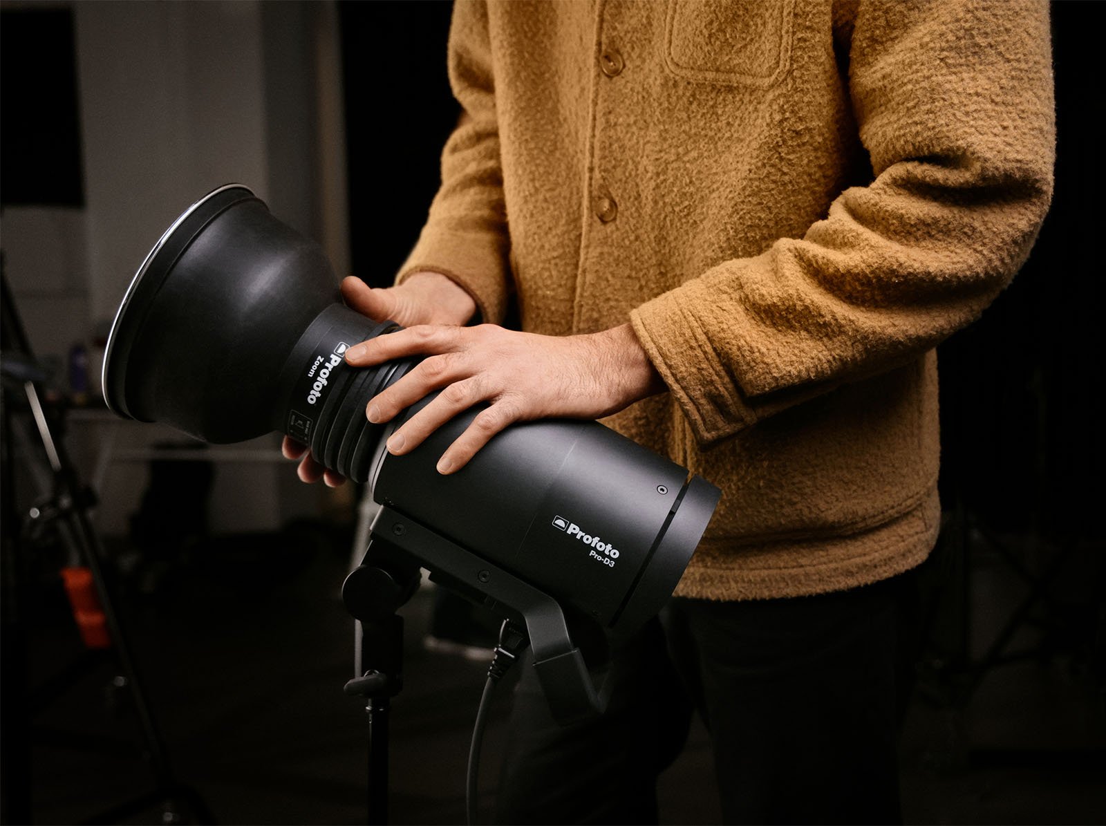 A person in a beige cardigan adjusts a professional studio light, focusing on its angling. details are visible in a dimly lit photography studio setting.