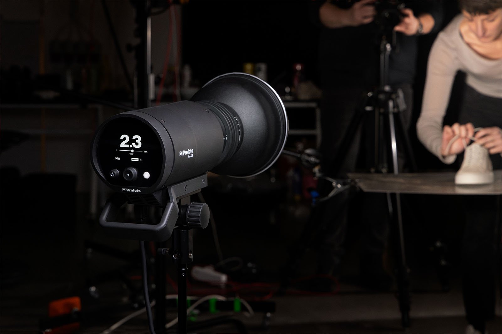 A professional studio setting showing a close-up of a profoto studio light with a digital display reading "2.3" and a photographer working with a model blurred in the background.