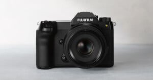 Front view of a black Fujifilm professional digital camera placed on a light gray surface. The camera features a prominent lens and detailed buttons on the left side of its body. The brand name "Fujifilm" is clearly visible on the top of the camera.