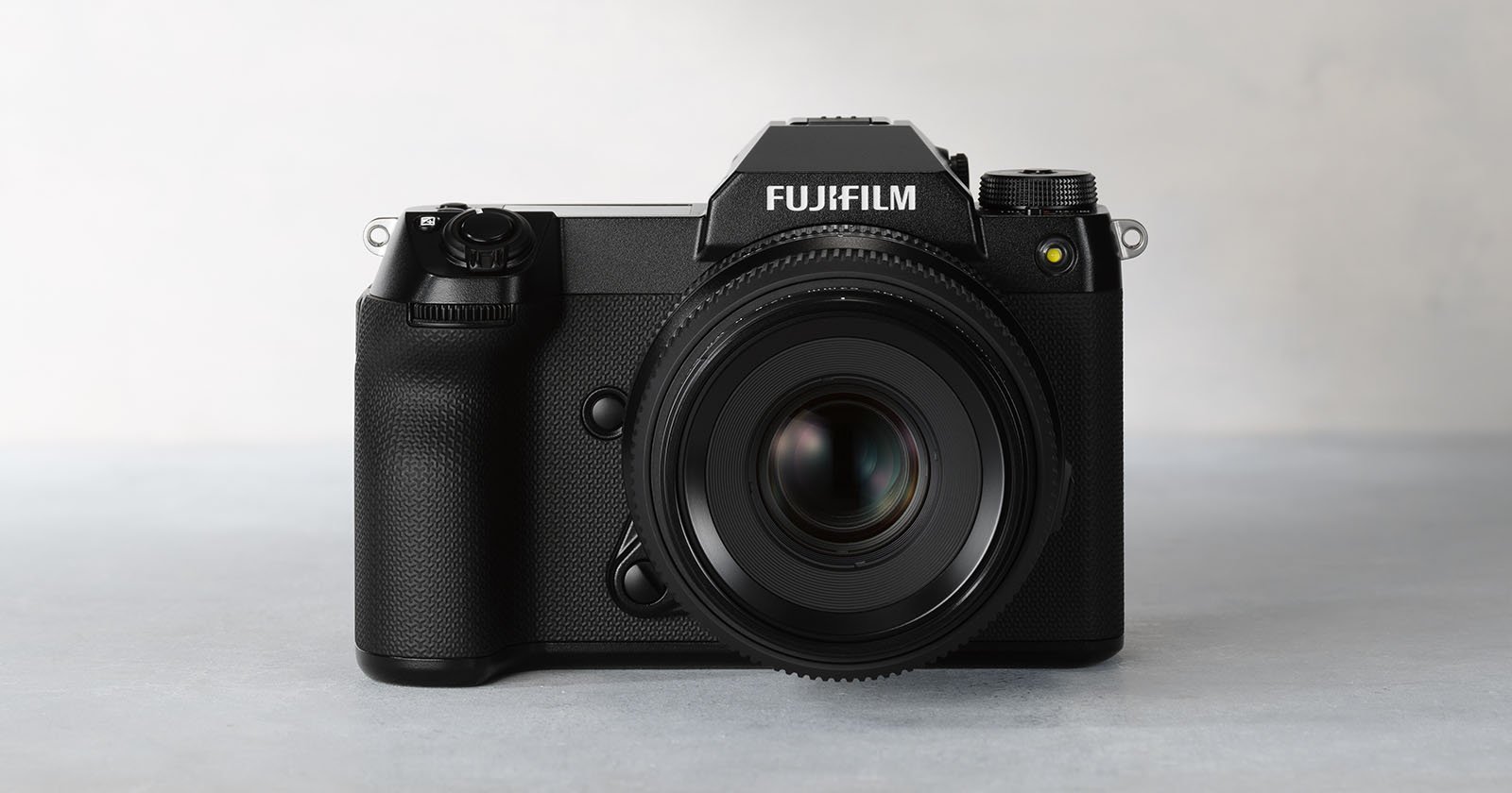 Front view of a black Fujifilm professional digital camera placed on a light gray surface. The camera features a prominent lens and detailed buttons on the left side of its body. The brand name "Fujifilm" is clearly visible on the top of the camera.
