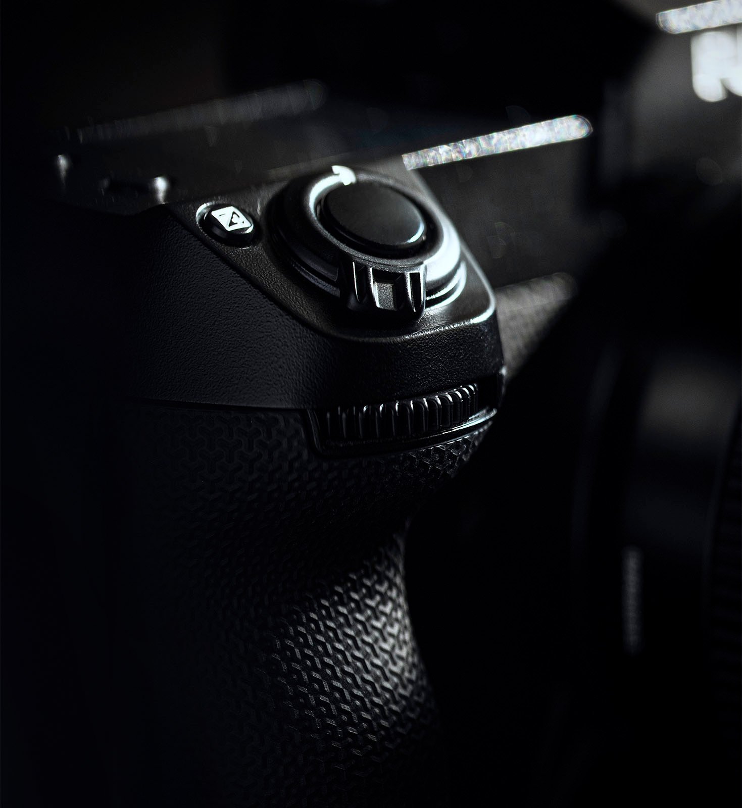 A close-up shot of a black camera's control dial and textured grip on a dark background. The image focuses on the intricate details of the dial and the ergonomic design of the grip.