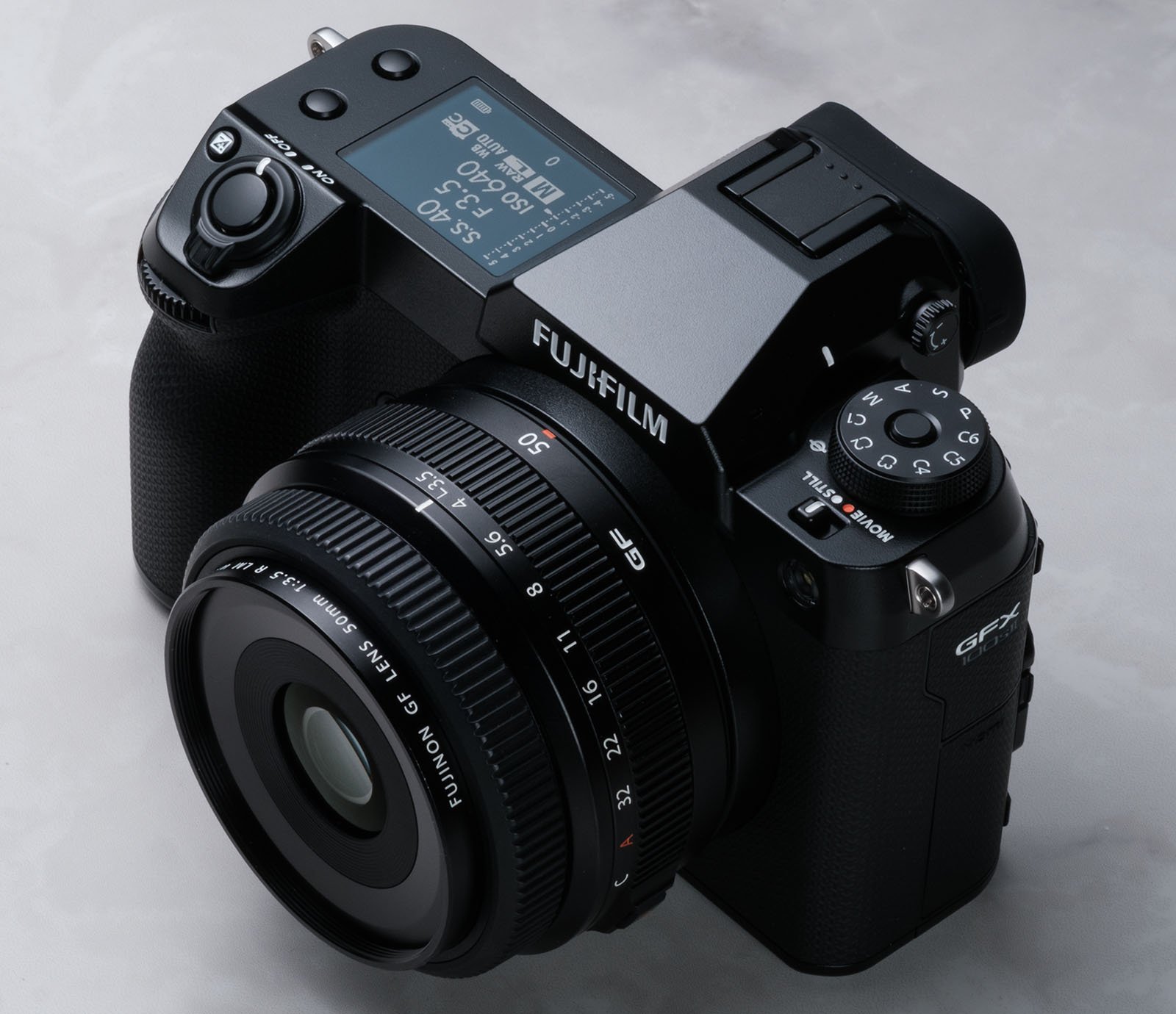 A Fujifilm GFX digital camera is shown. The camera features a large lens with various settings marked, a top display screen, and multiple buttons and dials for different camera functions. The body is black and has a textured grip. The camera rests on a light surface.