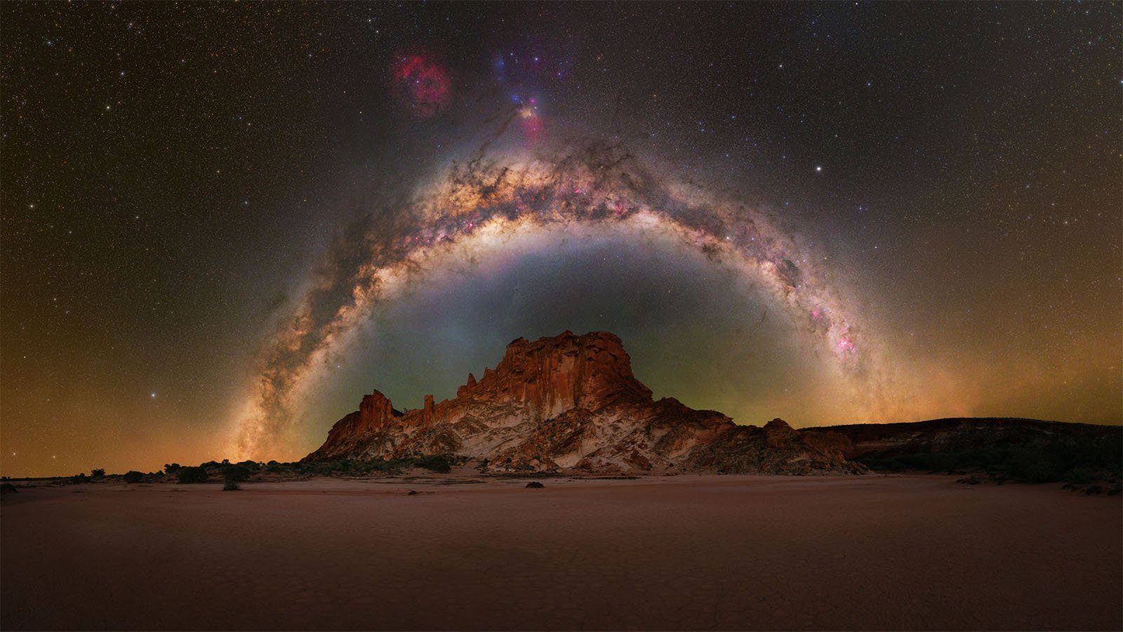  A mesmerizing night sky showcases a vividly colorful Milky Way arching over a rugged, rocky landscape. The terrain features scattered brush and a prominent cliff formation, illuminated by the brilliant starlight against a clear, dark sky.