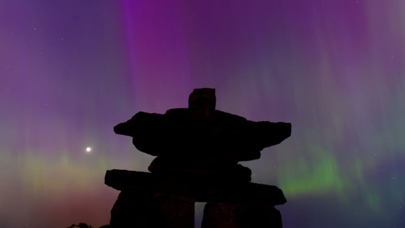 An inuksuk silhouette against a night sky illuminated by vibrant purple and green aurora borealis, with a bright star visible near the horizon.