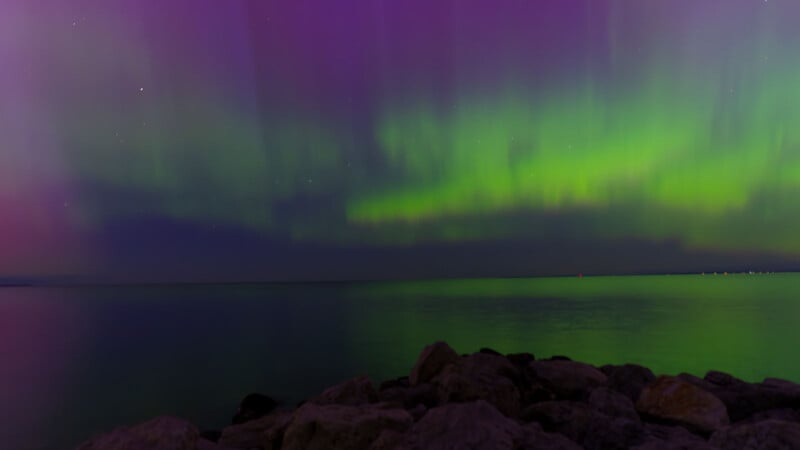 Northern lights displaying vibrant green hues over a calm sea with a rocky shoreline in the foreground under a starlit sky.