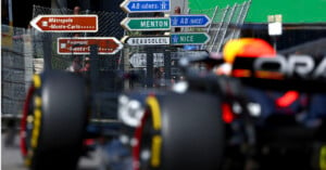 A Formula 1 race car speeds past a series of road signs indicating directions to various destinations, including Monte-Carlo, Menton, Nice, and Beausoleil. The backdrop includes a wire fence and a clear blue sky.