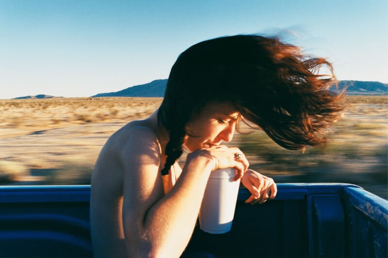 A person with long, wind-swept hair sits in the back of a moving vehicle, holding a cup with both hands. The background features a desert landscape and clear blue skies.
