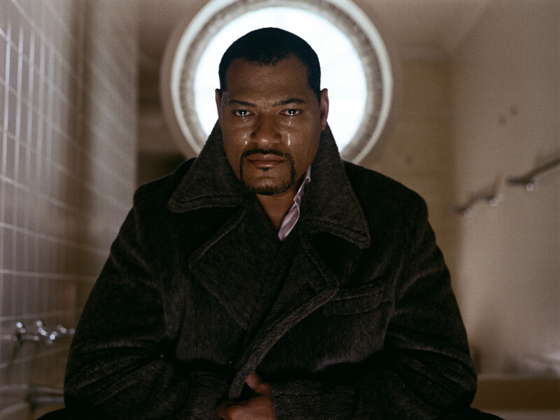 A man with a solemn expression is sitting indoors, wearing a dark coat. Light from a circular window behind him illuminates his face, highlighting the tear tracks on his cheeks. The background features tiled walls and blurred details, emphasizing his emotional state.