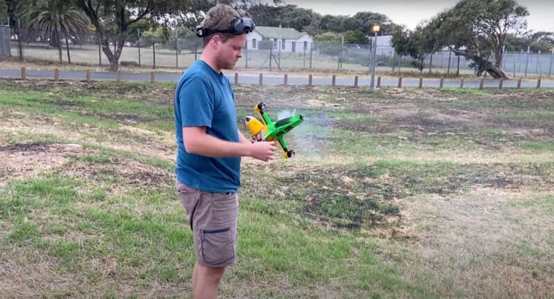 A man stands in a field wearing fpv (first-person view) goggles and holding a green and yellow toy airplane, with a light cloud of smoke visible near the plane’s rear.
