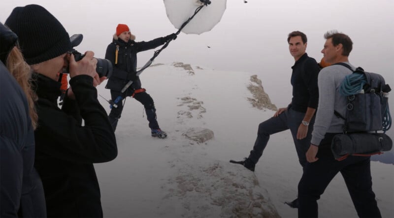 Photographer takes a picture of two men posing on a snowy mountain peak. Another person holds a large reflector above the subjects, aiding in lighting for the shot. All individuals are dressed in warm clothing suitable for cold weather. The background is overcast.