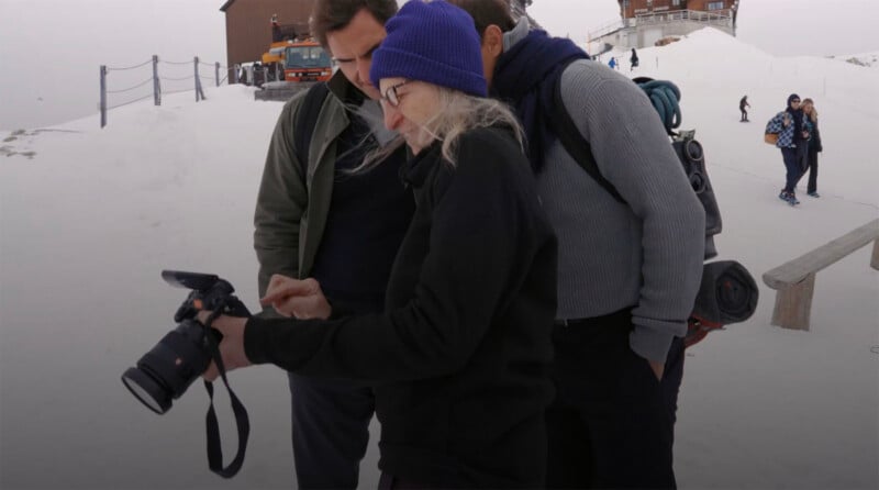 Three people stand on snow-covered ground, closely looking at a camera screen. One person, wearing a purple beanie and glasses, holds the camera. A building, a parked truck, and other people walking are visible in the background.