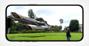 A smartphone screen displaying an augmented reality scene of a futuristic spacecraft hovering over a grassy park. A person is standing in the park looking up at the spacecraft. Trees and a building are visible in the background.