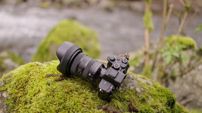 A professional camera with a large lens resting on a moss-covered rock in a natural, outdoor setting, with a soft-focus background featuring a stream and foliage.