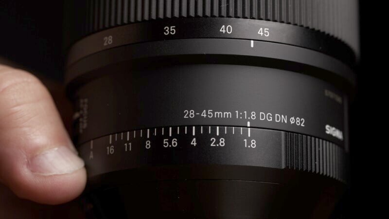 Close-up image of a person's hand adjusting the zoom ring on a camera lens. The lens markings indicate it is a 28-45mm f/1.8 model, with various aperture settings visible. The lens is black and the background is out of focus.