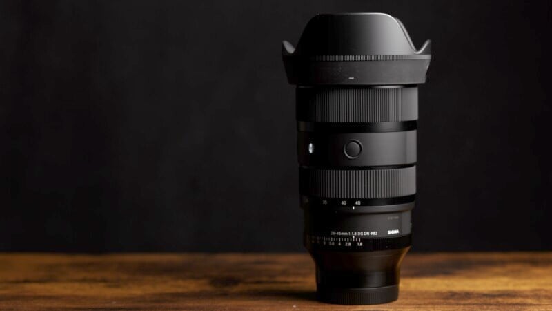 Photo of a black Sigma camera lens with lens hood, placed vertically on a wooden surface against a dark background. The lens has multiple adjustment rings and a visible brand mark.