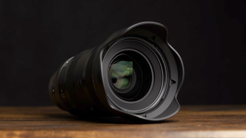 A close-up view of a DSLR camera lens with a lens hood attached, resting on a wooden surface. The background is dark, focusing attention on the lens's glass elements, which slightly reflect light green hues.
