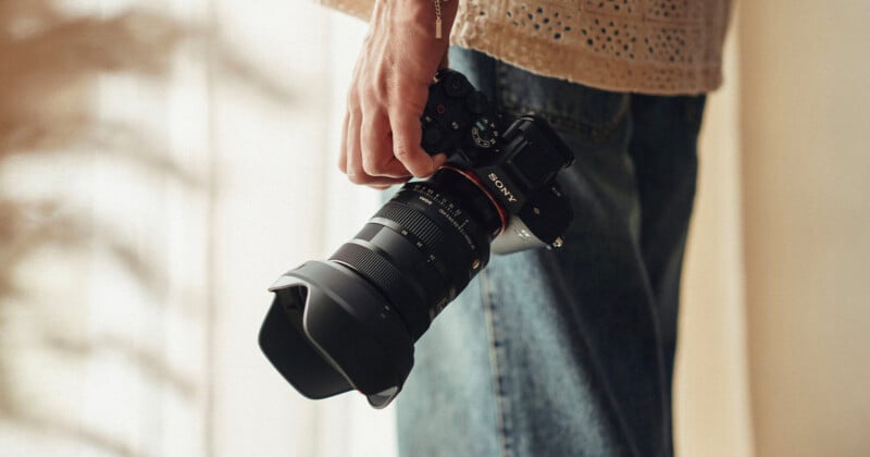 A close-up of a person holding a Sony camera with a large lens attached. The individual is wearing a beige, textured top and blue jeans, with the camera hanging by their side. The background is softly lit with blurred, neutral tones.