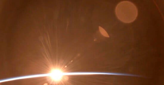 A striking view of a bright light source at the edge of earth’s atmosphere, illuminating flares and casting a warm glow with lens flares and bokeh effects apparent.