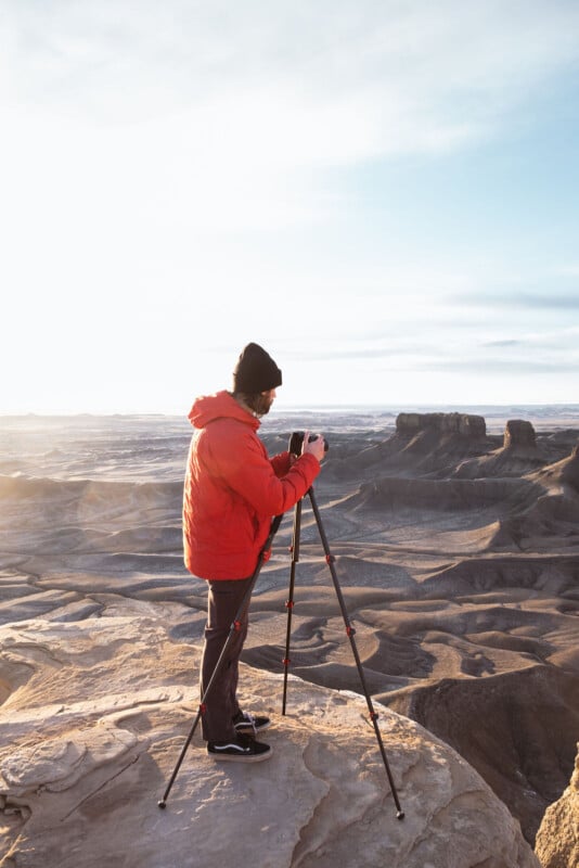 A person in a bright red jacket and black beanie stands on a rocky ledge overlooking a vast desert landscape with tripod and camera setup. The sky is clear, and the sun casts a warm light on the scene, highlighting the rugged terrain below.